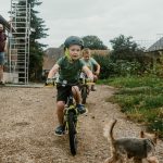 Two kids cycling on a dirt path with a small dog chasing them, adult supervising nearby.
