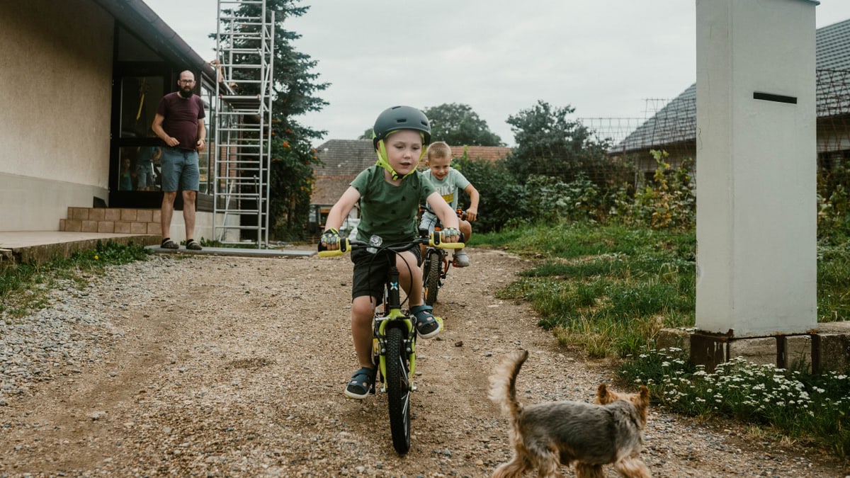 Two kids cycling on a dirt path with a small dog chasing them, adult supervising nearby.